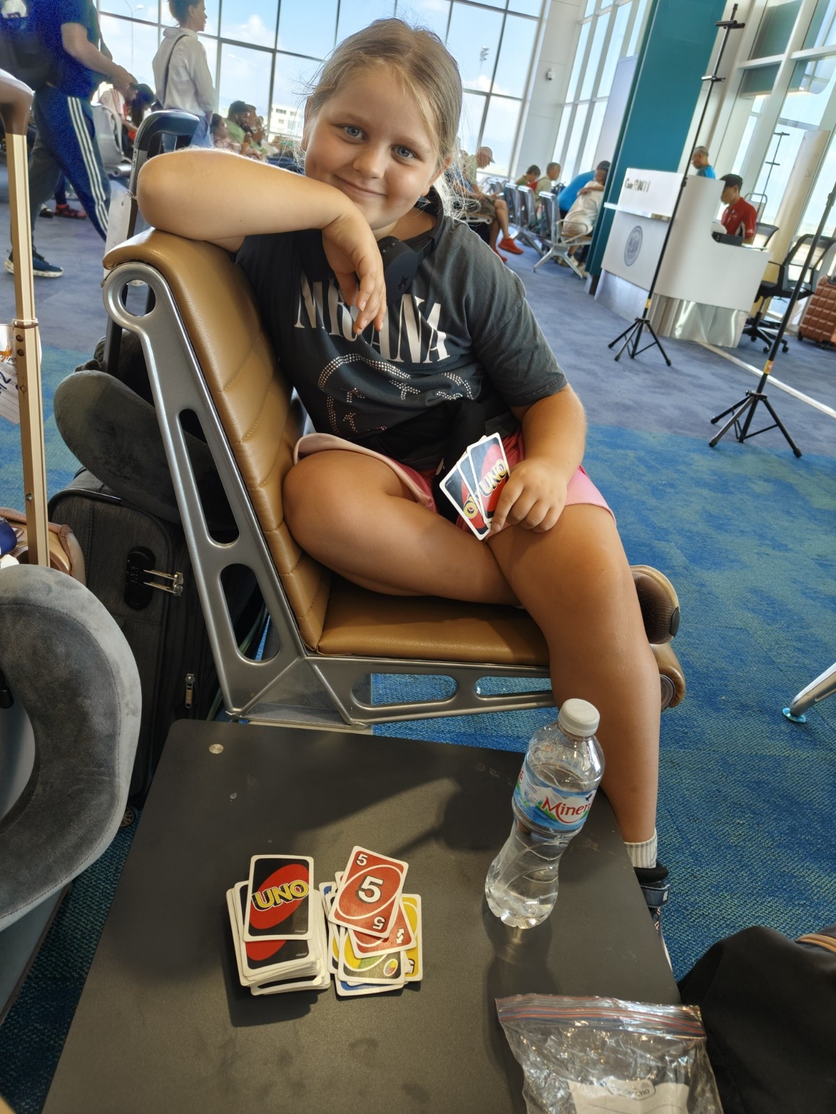 Deklica sedi na letališču in igra karte UNO ali Enka.