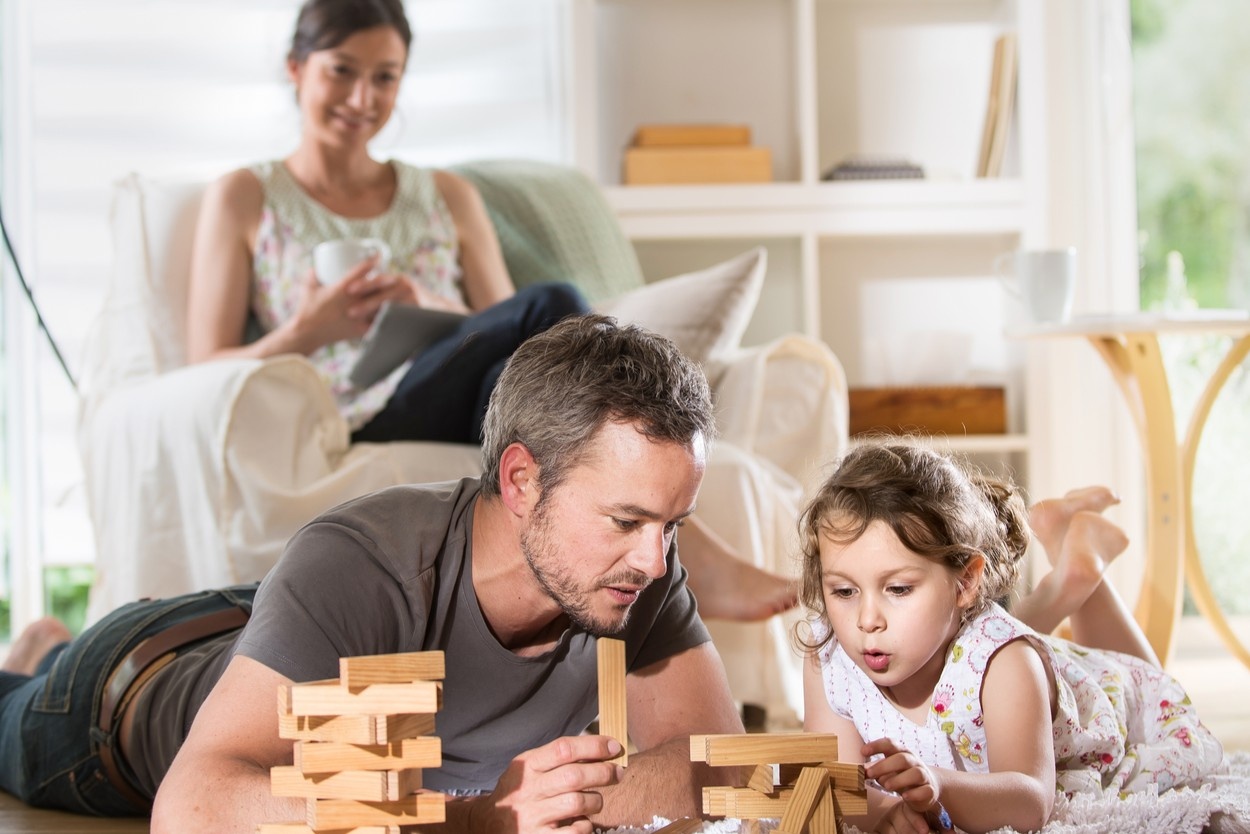 Oče in hčerka se igrata jenga stolp, v ozadju ju gleda mama.