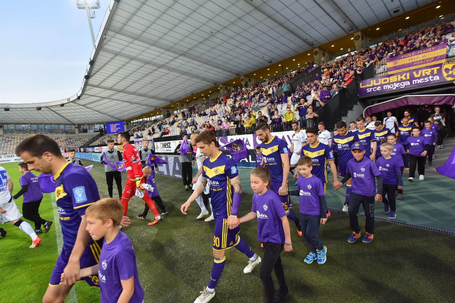 Nogometaši v vijoličnih dresi hodijo na igrišče in za roke držijo otroke, ki so tudi oblečeni v vijolične majice.