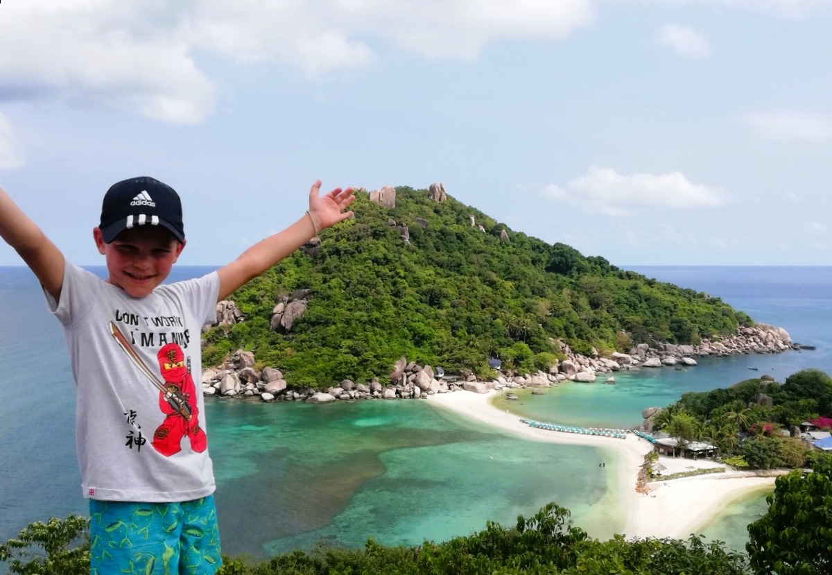 Otrok, oblečen v sivo majico z napisom in modre hlače, stoji in z razprtimi rokami kaže na tropski otok za njim
