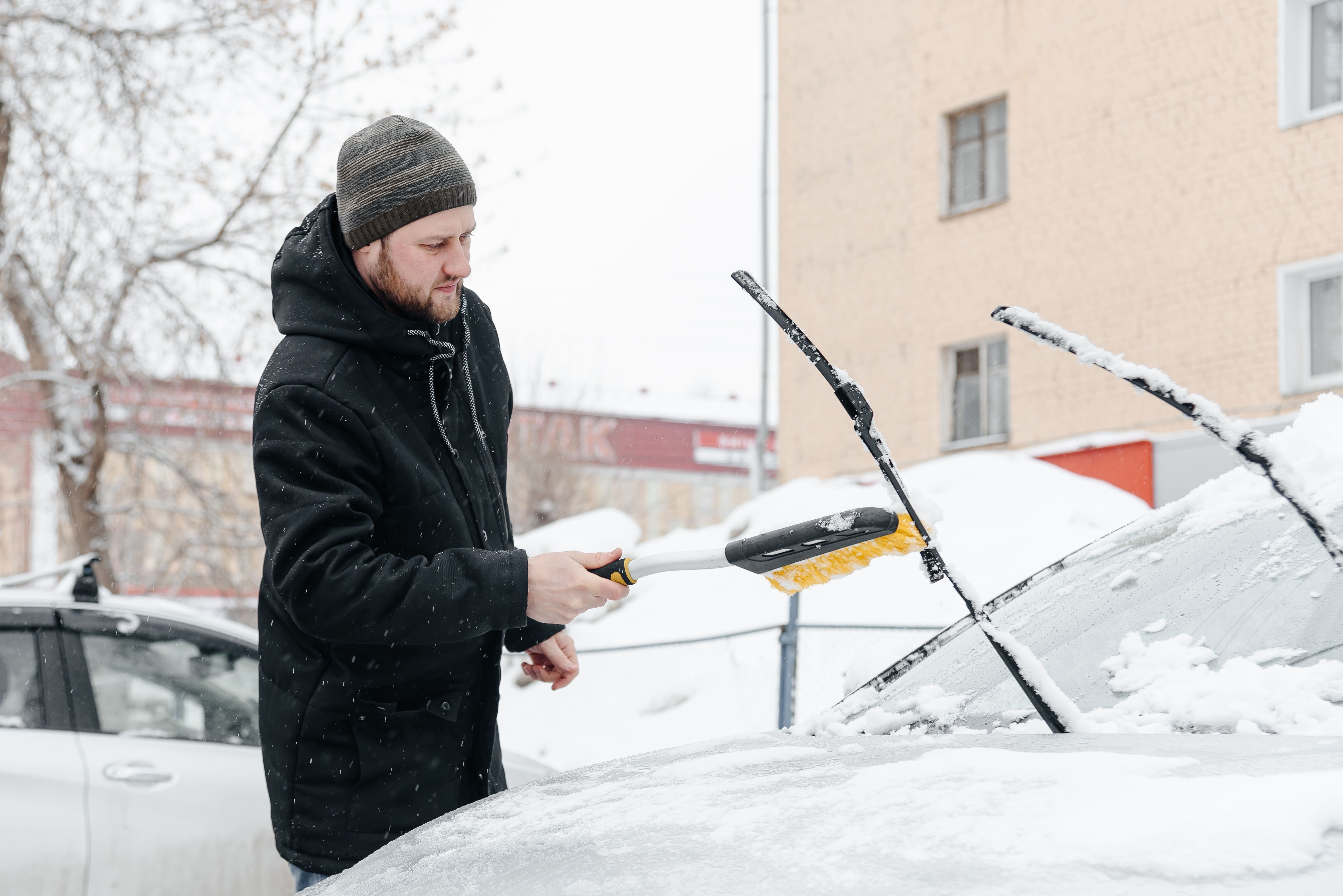Moški, ki je oblečen v zimsko bundo, čisti brisalce in vetrobransko steklo z metlico za sneg.