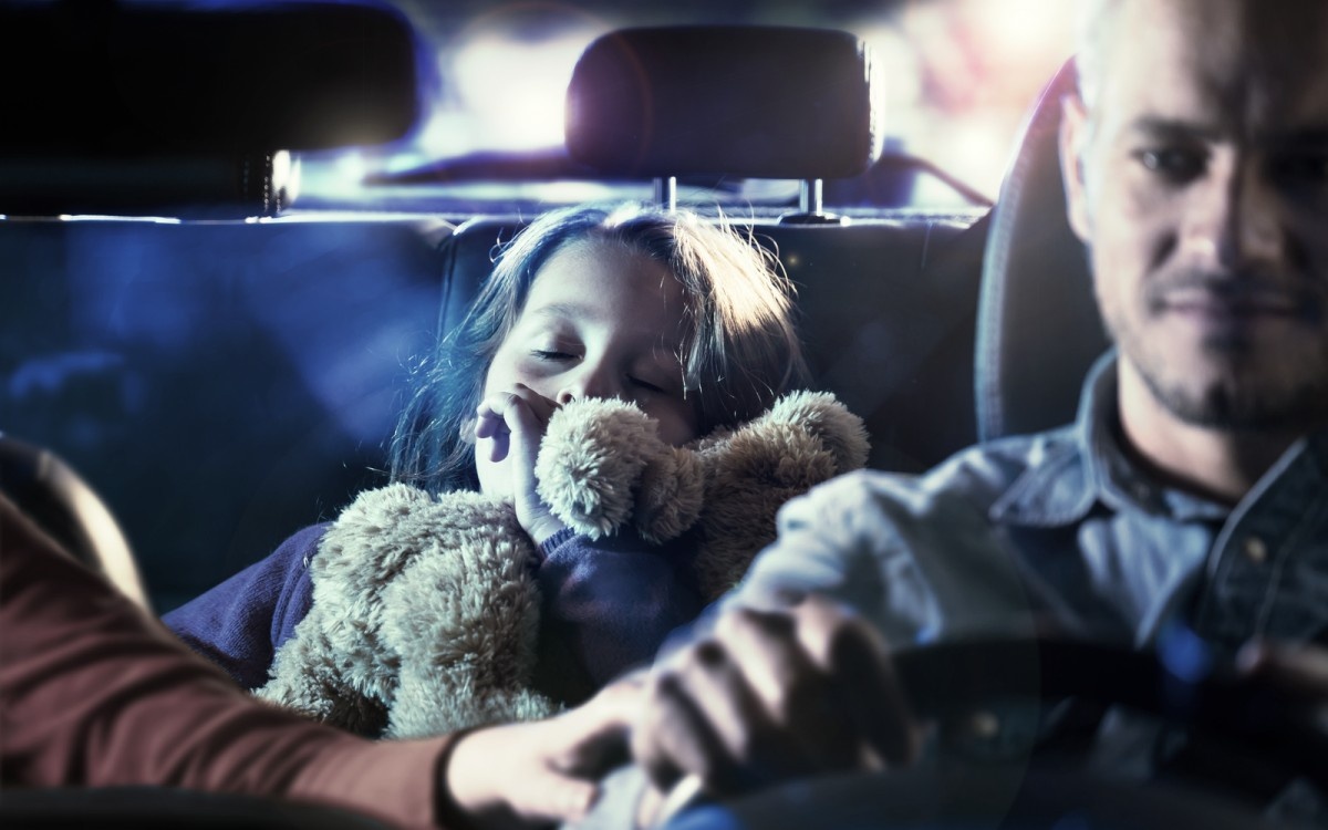 Moški vozi avto, za njim spi otrok z igračo v rokah.