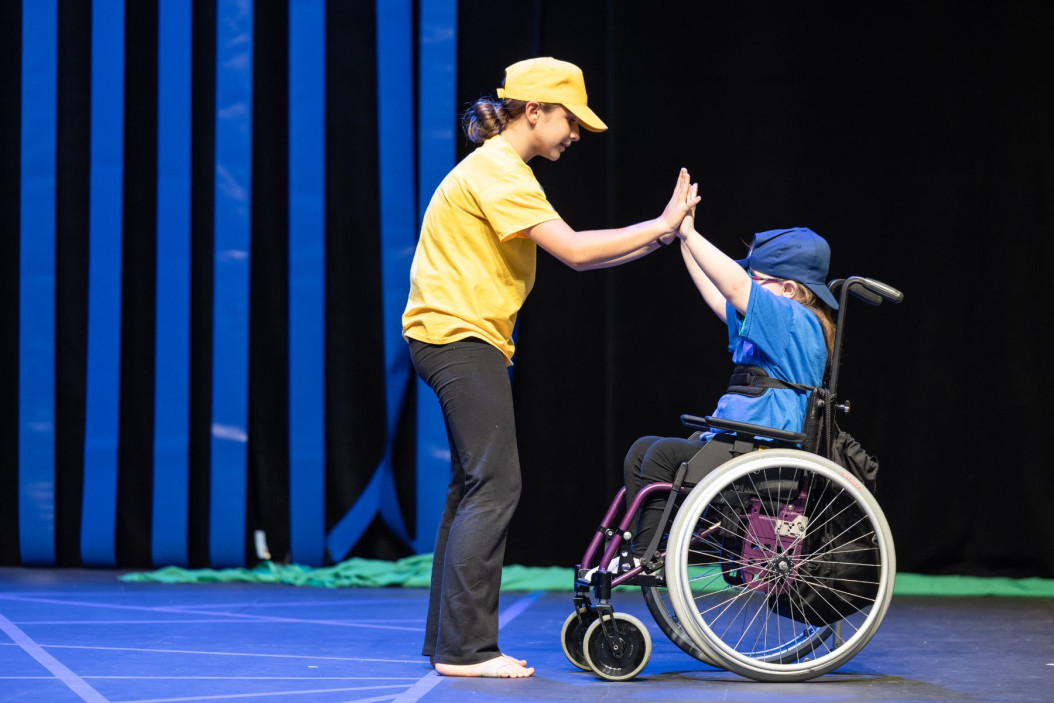 Fotografija dveh otrok sredi plesa, eden je oblečen v rumeno majico, drugi je na vozičku in oblečen v modro majico.