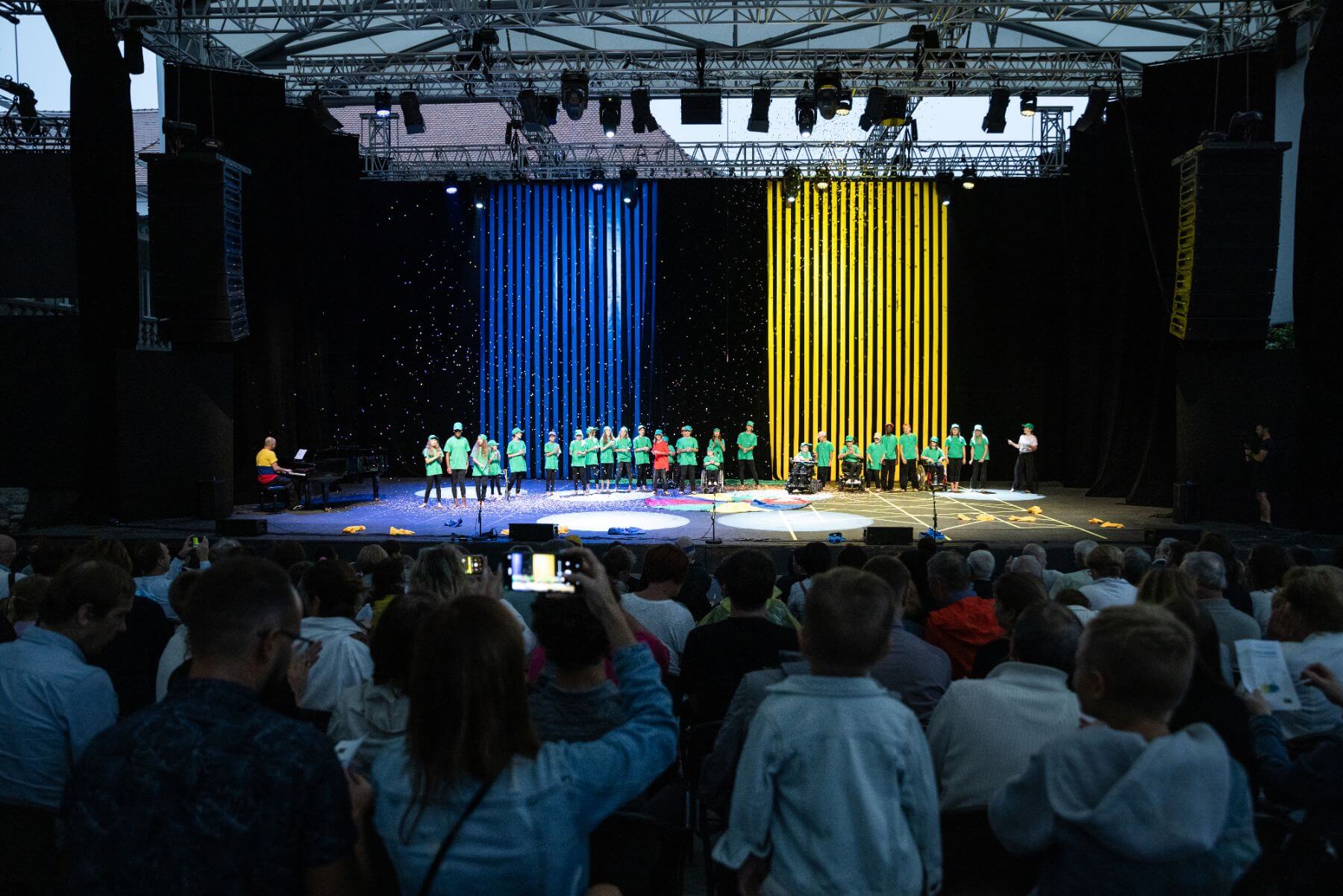 Otroci v zelenih majicah na koncu nastopa, v ozadju so modri in rumeni trakovi.