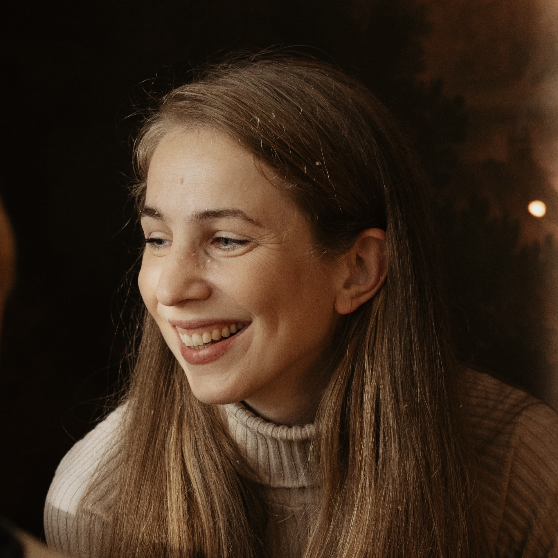 Mlada ženska z dolgimi ravnimi rjavimi lasmi se smeji, oblečena je v bež pleten pulover