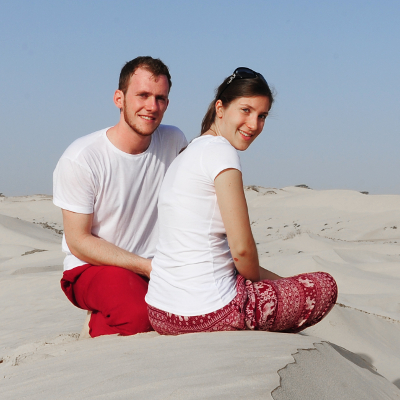 Fotografija moškega in ženske, oblečena sta v beli majici in rdeče hlače, sedita sredi puščave in gledata proti kameri
