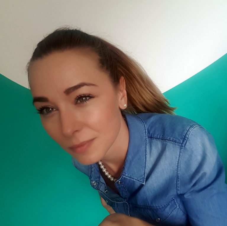 Ženska s svetlo rjavimi lasmi v čopu, oblečena je v modro srajco in stoji pred belo zeleno steno