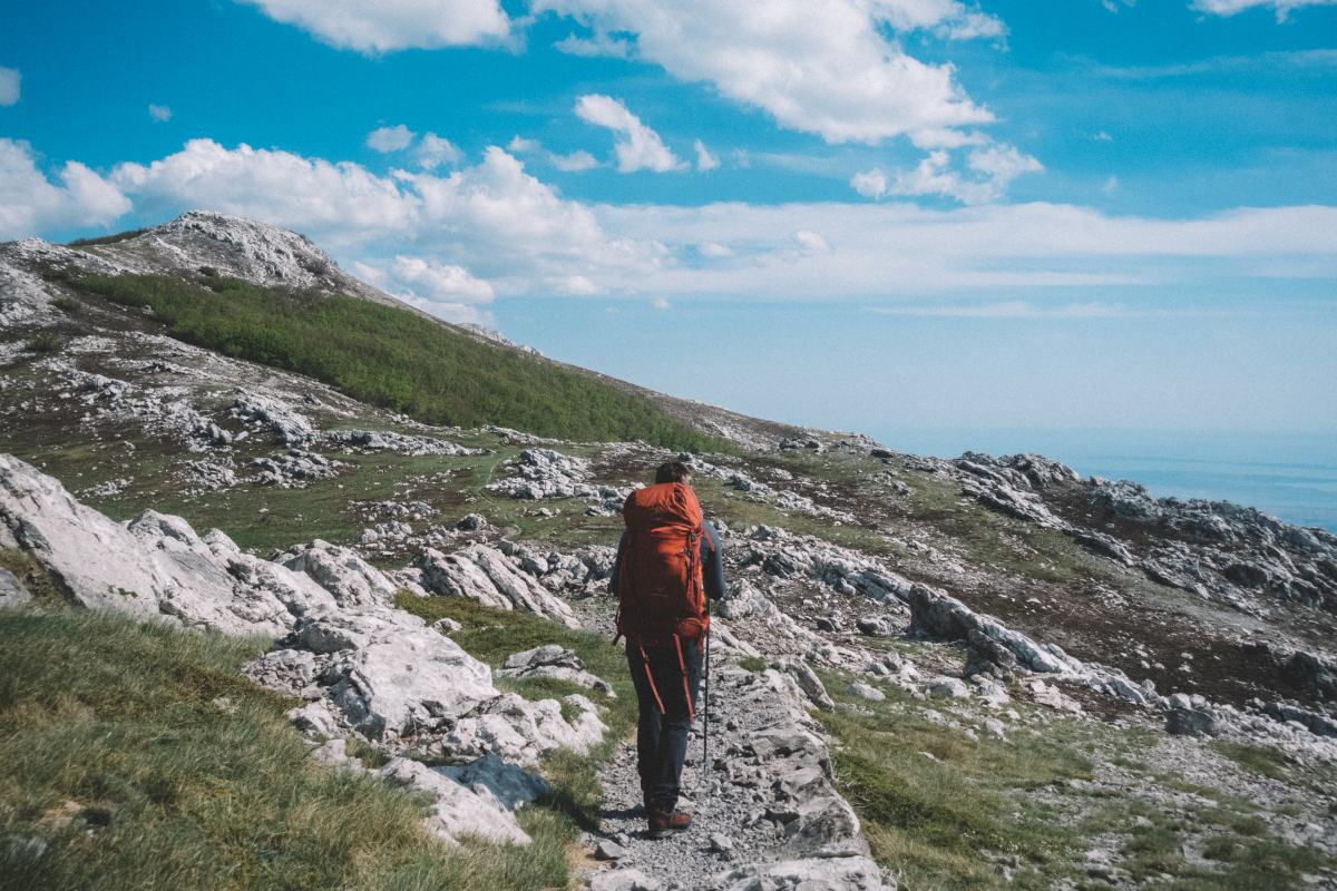 Moški z velikim oranžnim nahrbtniku hodi po poti na hribu.