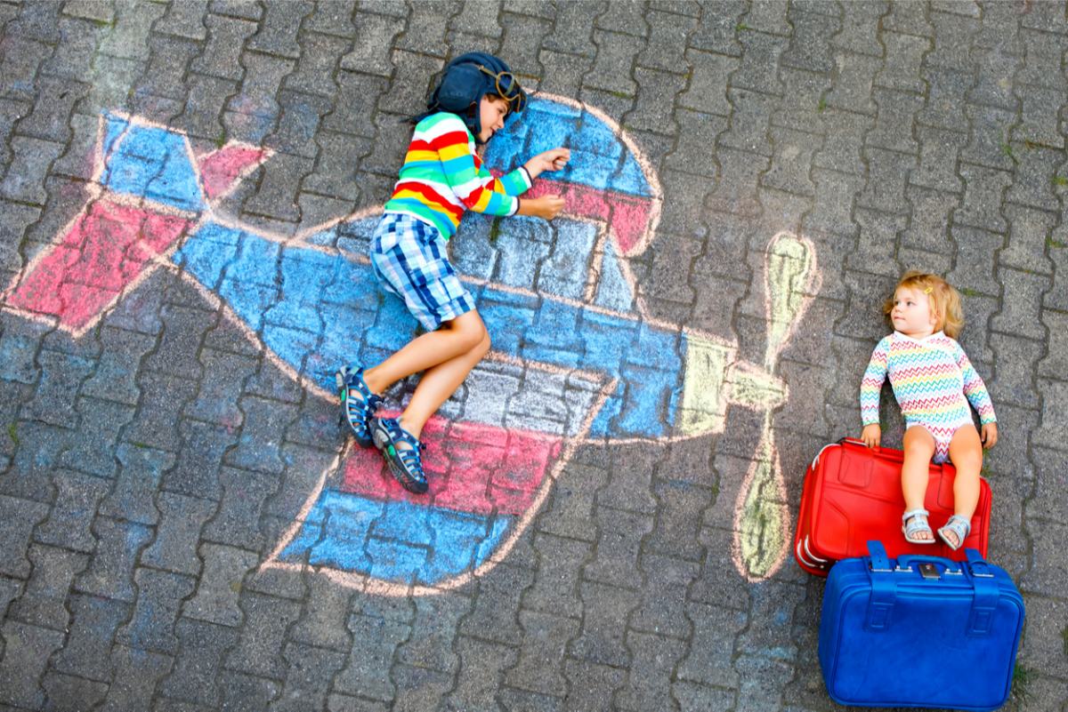 Dva otroka ležita na risbi narisani s kredami, fantek leži na letalu, punčka pa ja naslonjena na potovalke.