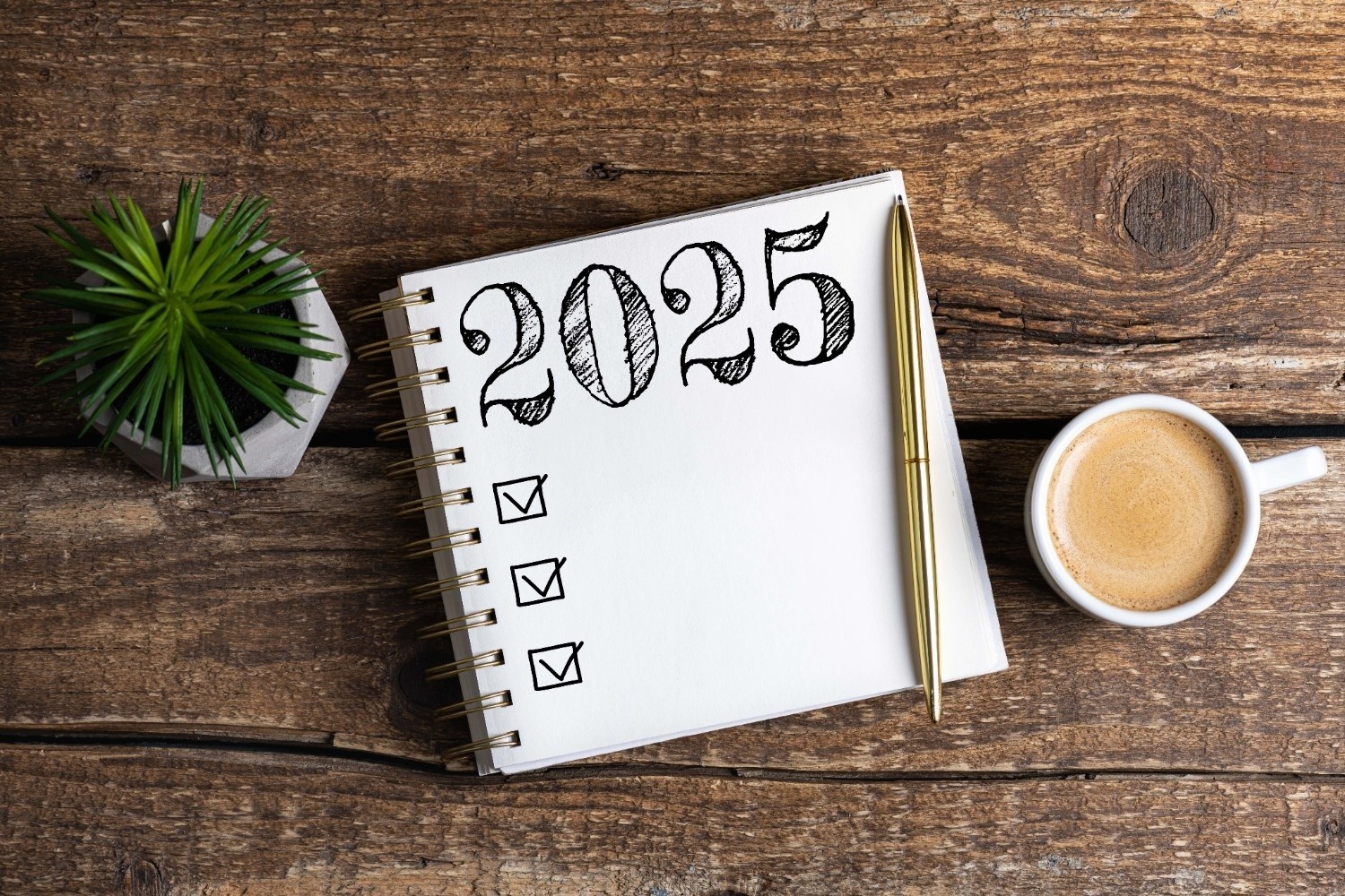 Beležka z napisom 2025, zraven je svinčnik, skodelica kave in majhna rastlina.
