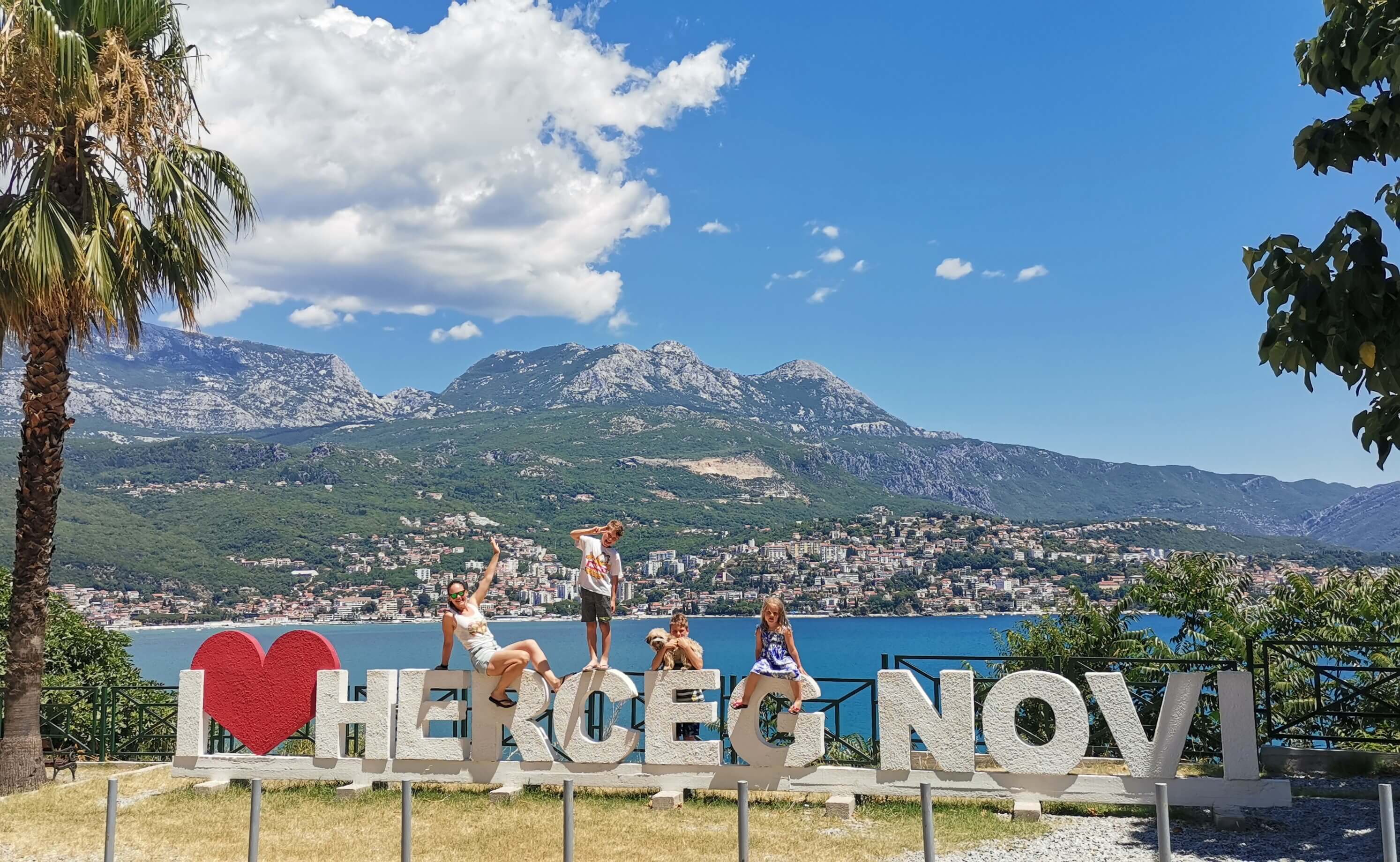 Mlada družina sedi na napisu I <3 Herceg Novi pred obalnim mestecem.