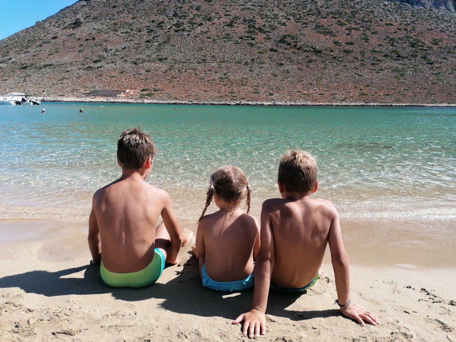 trije otroci sedijo na plaži in gledajo morje.