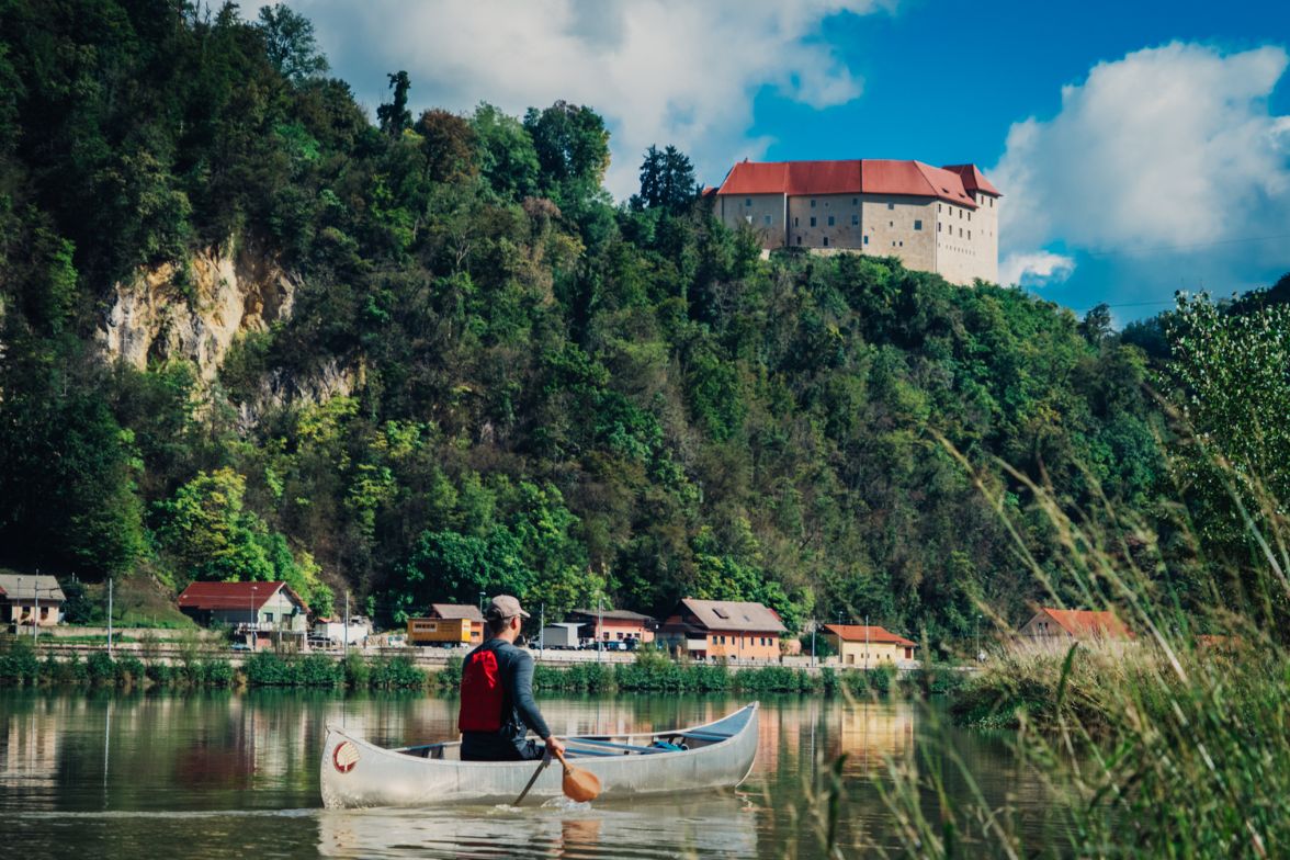 Oseba na kajaku vesla po reki, v daljavi se vidi grad na vrhu hriba.