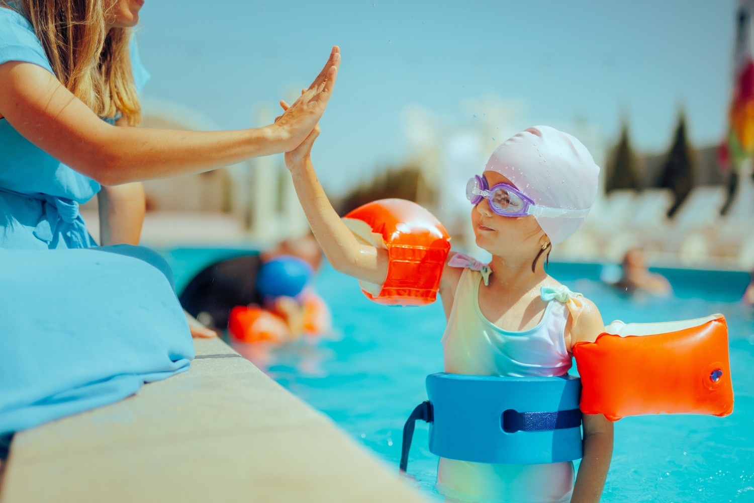 Otrok s plavalnimi rokavčki stoji v bazenu in samozavestno daje petko ženski, ki sedi na robu bazena.