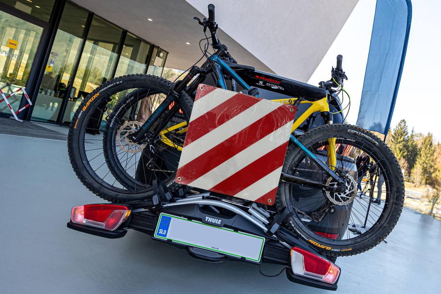 Dve gorski kolesi sta nameščeni na zadnjem nosilcu za kolesa, pritrjenem na avtomobil, za kolesoma pa je nameščena rdeče-bela opozorilna tabla.